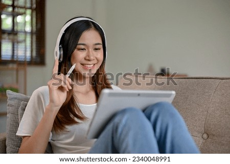 Young cheerful Asian girl using digital tablet in her free time while sitting on the sofa in the living room. 