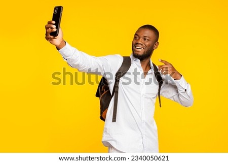 Young male student of african american ethnicity wears white shirt and backpack, doing selfie shot on mobile phone post photo on social network pointing finger at camera isolated on yellow background.