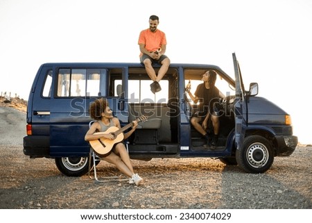 A group of 3 young people from all walks of life are having fun in a camper van parked on a mountain. The woman with the afro hair plays a flamenco guitar while her friends look at each other. Royalty-Free Stock Photo #2340074029