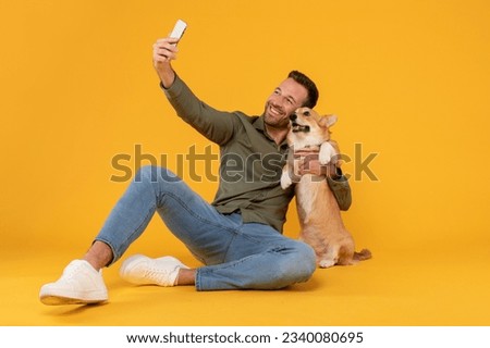Excited european man taking selfie with his pretty corgi dog, posing looking at phone camera embracing pet, isolated on yellow studio background wall Royalty-Free Stock Photo #2340080695