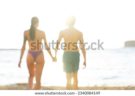 Blurred photo of handsome man wearing swimsuit with beautiful woman walks on the beach in a sunny day, holding hands, during holidays. Young adults caucasian couple travel together a romantic vacation