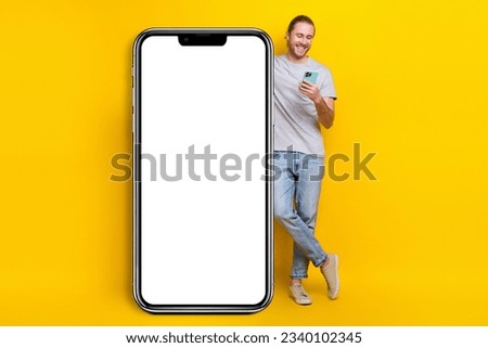 Full size photo of cheerful man gray t-shirt denim pants standing near white banner look at smartphone isolated on yellow color background