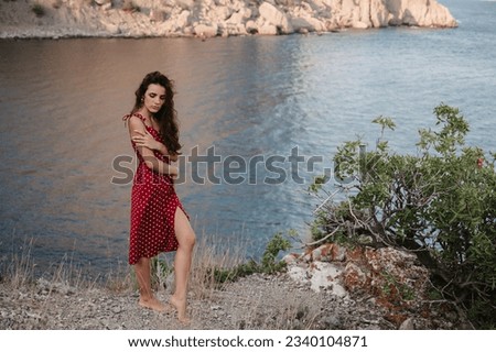 A picture of a young, attractive Caucasian woman in a red dress at the edge of a cliff near the beach and the sea