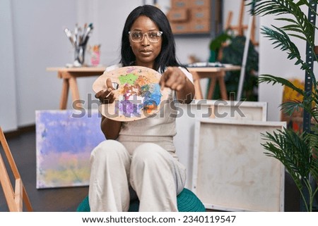 African young woman holding painter palette pointing with finger to the camera and to you, confident gesture looking serious 