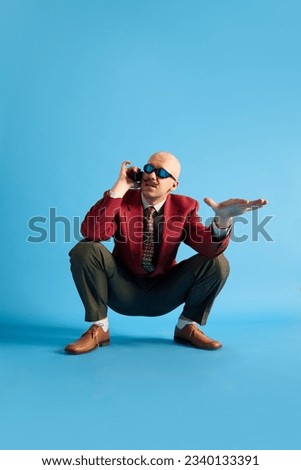 Balded man with mustache wear in vintage style squatting and talking on retro phone looking away over blue background. Concept of fashion, emotions, communication, vintage, retro and ad Royalty-Free Stock Photo #2340133391