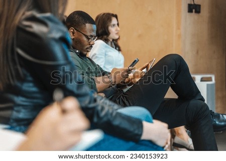 Side view photo of black male employee checking his phone while holding a tablet. Employee not paying attention to a presentation Royalty-Free Stock Photo #2340153923