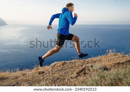 male runner running uphill trail on edge in blue jacket and black tights, background of sky and sea Royalty-Free Stock Photo #2340192513
