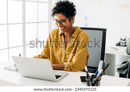 Graphic designer smiles as she works on a laptop in an office. Woman using a graphics tablet to make drawing designs. Creative business woman enjoys working on her project in an office. Royalty-Free Stock Photo #2340195125