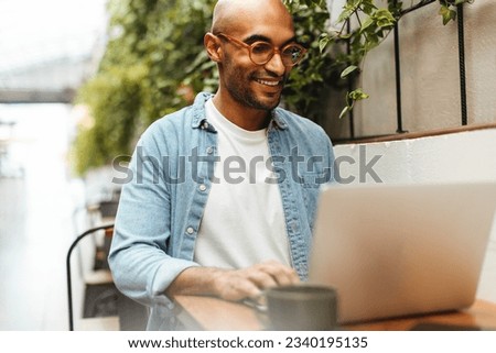 Young black man smiling as he freelances with his laptop at a coworking space, enjoying the ability to work remotely using internet connection. Male digital nomad sitting in a coffee shop. Royalty-Free Stock Photo #2340195135