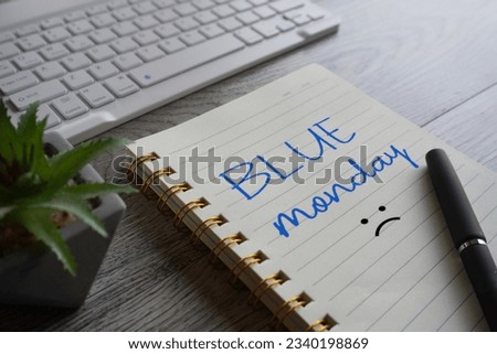 Close up image of notebook with text BLUE MONDAY, pen and laptop on table