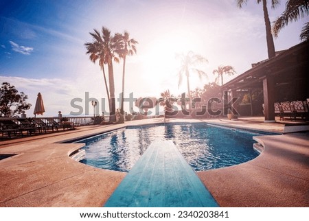 View from behind of a diving board to swimming pool, sunbeds and umbrellas lit with warm sunset light going though leaves of palm tree Royalty-Free Stock Photo #2340203841