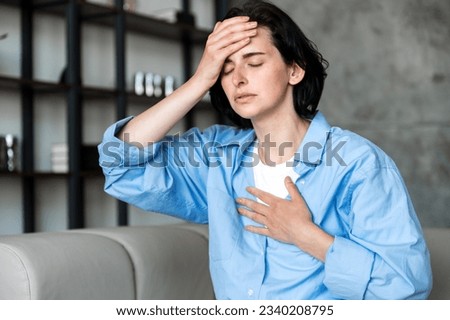 Female having heart, asthma or panic attack. Difficulties with breathe, feeling severe pain. Woman sitting on sofa with closed eyes at home and holding hands on chest and forehead. Royalty-Free Stock Photo #2340208795