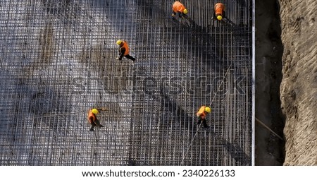 Construction site. Workers in orange uniform carrying steel bars. Rebars floor, builders working around. Top view, daylight. High quality photo