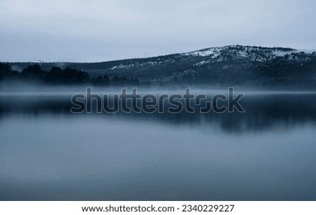 Long exposure shot of the lake at sunrise. Panorama view of the mountains and forest reflection in the water surface in an early morning with fog.  Royalty-Free Stock Photo #2340229227
