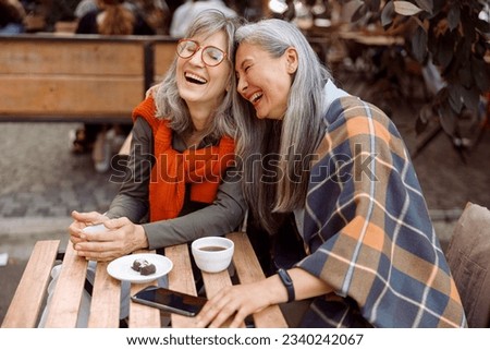 Pretty senior Asian woman puts head on laughing friend shoulder resting together at table in street cafe on nice autumn day Royalty-Free Stock Photo #2340242067