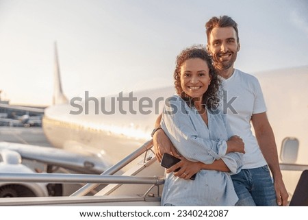 Portrait of happy couple of tourists, man and woman looking excited while standing together outdoors ready for boarding the plane at sunset. Vacation, lifestyle, traveling concept