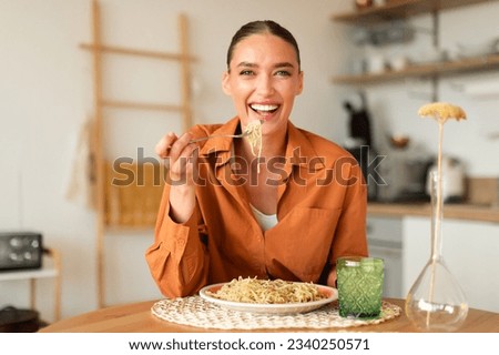 Happy young european lady eating delicious homemade Italian pasta, enjoying tasty lunch, looking at camera and laughing, sitting in cozy kitchen interior, copy space Royalty-Free Stock Photo #2340250571