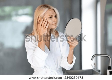 Skin Aging. Attractive Mature Woman Holding Mirror And Looking At Her Wrinkles Near Eyes, Beautiful Senior Female Wearing Bathrobe Checking Face, Noticing Age Changes, Standing In Bathroom Interior Royalty-Free Stock Photo #2340250673