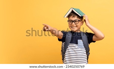 Excited cheerful positive school aged kid boy wearing eyeglasses with book on his head and backpack pointing at copy space for text or advertisement, isolated on yellow background, panorama Royalty-Free Stock Photo #2340250769