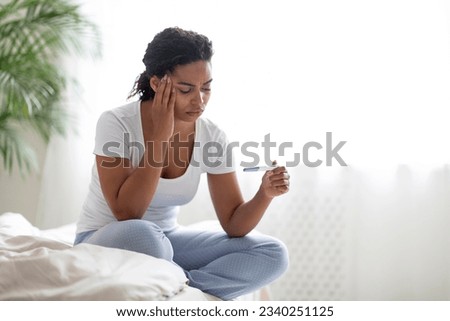 Unhappy sad young african american woman with pregnancy test in hand sitting on bed in white bedroom interior, upset frustrated black lady suffering infertility or thinking about unwanted pregnancy Royalty-Free Stock Photo #2340251125
