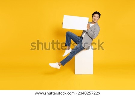 Young Asian man sitting and showing blank white billboard isolated on yellow background
