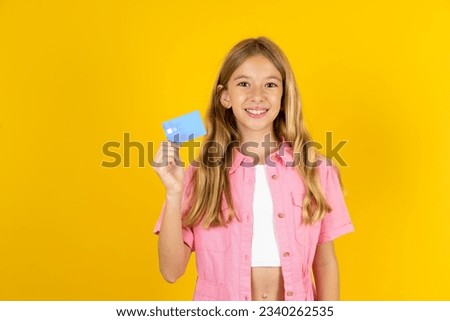 Photo of happy cheerful smiling positive Caucasian kid girl wearing pink jacket over yellow background recommend credit card