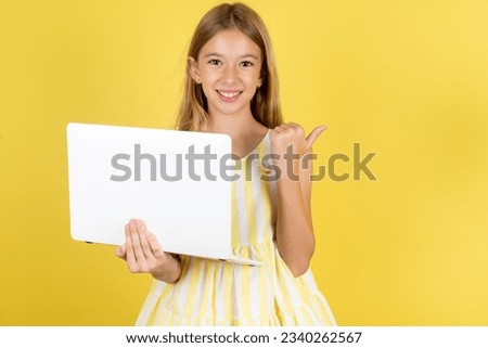 Caucasian kid girl wearing yellow dress over yellow background directing empty space  hold laptop