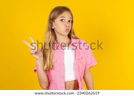 Caucasian kid girl wearing pink jacket over yellow background makes peace gesture keeps lips folded shows v sign. Body language concept
