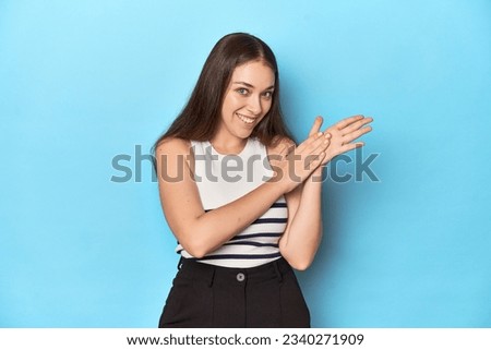 Woman in a striped top posing on a blue studio backdrop feeling energetic and comfortable, rubbing hands confident. Royalty-Free Stock Photo #2340271909