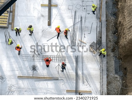 Workers in orange and yellow uniform working with reinforcement at the construction site, gray concrete background. Stack of bars and constructions around. High quality photo
