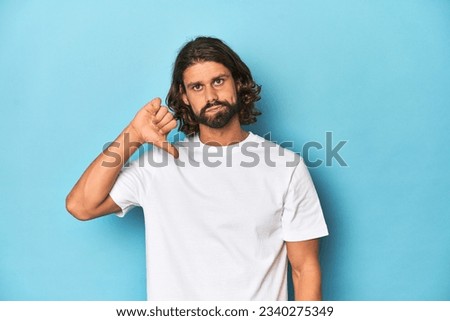 Bearded man in a white shirt, blue backdrop showing thumb down, disappointment concept. Royalty-Free Stock Photo #2340275349