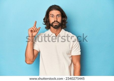 Long-haired man in a white polo, blue studio having an idea, inspiration concept. Royalty-Free Stock Photo #2340275445