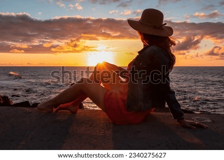 Beautiful luxury woman in summer hat and red dress sitting at port of Puerto de la Cruz during sunset, Tenerife, Canary Islands, Spain, Europe. Vacation at the sea. Reflection of sun beams in the sea
