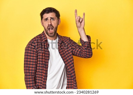 Caucasian man in red checkered shirt, yellow backdrop showing a horns gesture as a revolution concept.