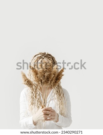 Woman holding pampas grass in front of face, light background, autumn, fall minimal trend concept. Female with long hair dreadlocks hiding behind bouquet dried plant. Creative portrait photo ideas