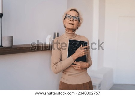 Reminiscing elderly lady in casual clothes and eyeglasses standing near shelf with photo frame folded to breast