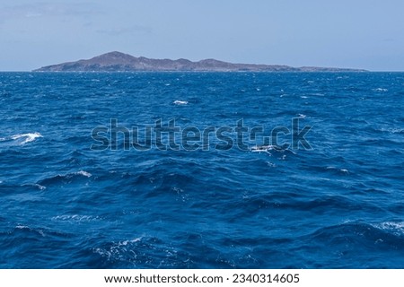 Small brown bare island rock without plants on it in the middle of the open blue ocean in the sunlight.