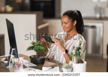 African female owner small business using smartphone taking photo bonsai for online sale while vlogging about plant care at home.