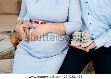 hands on the stomach of a pregnant woman in a gray dress with a red belt close-up male hands holding baby booties