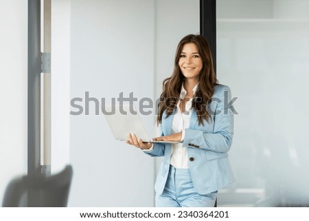 Portrait of beautiful smiling woman, successful manager holding laptop working online standing in modern office. Confident copywriter typing on keyboard. Remote job concept 