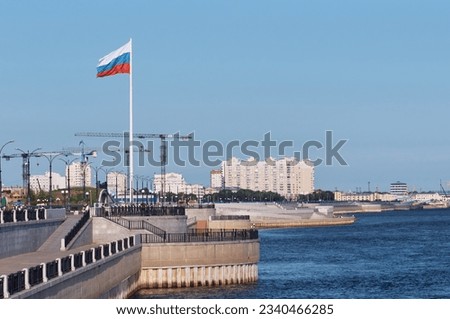 Large flag of Russia on the embankment of the Amur River in the city of Blagoveshchensk. Summer early morning on the border with China. Cranes at the construction site of the cable car terminal.