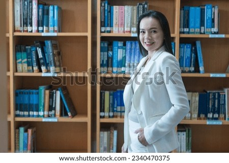 A female university lecturer in a business suit is searching for book on bookshelf in the library
