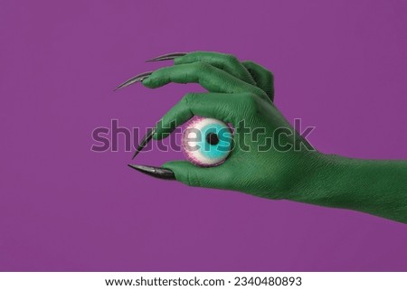 Green hand of witch with eye on purple background, closeup. Halloween celebration