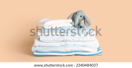 Stack of clean baby clothes and funny toy on beige background