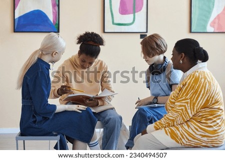 Diverse group of teenagers sitting in circle and holding books while discussing art lecture in modern art gallery or museum