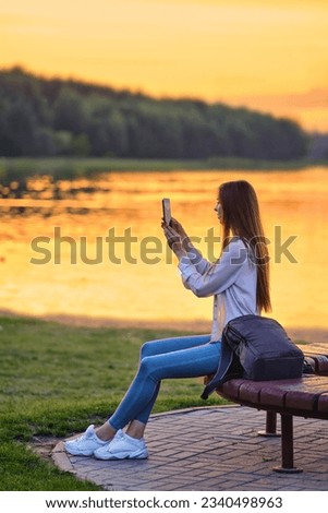 Young woman taking photos sitting on bank of river at sunset