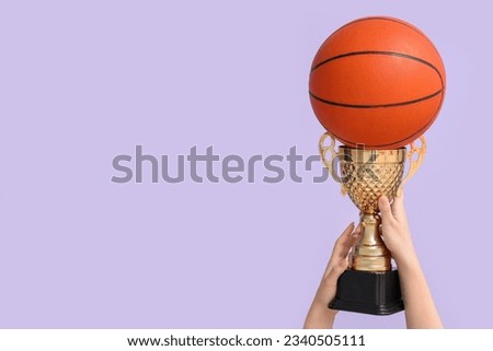 Female hands with gold cup and basketball on lilac background