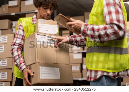 Male inventory manager checking parcel information on digital tablet and worker holding cardboard boxes. A stock of parcels with products ready for shipment in the background. Close-up