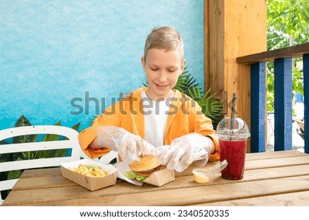 A cute boy in a street cafe eats fast food with lemonade in plastic disposable gloves in summer. Lunch outside, burger with fries