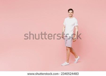 Full body side view young caucasian man wearing white t-shirt casual clothes walking going strolling looking camera isolated on plain pastel light pink background studio portrait. Lifestyle concept